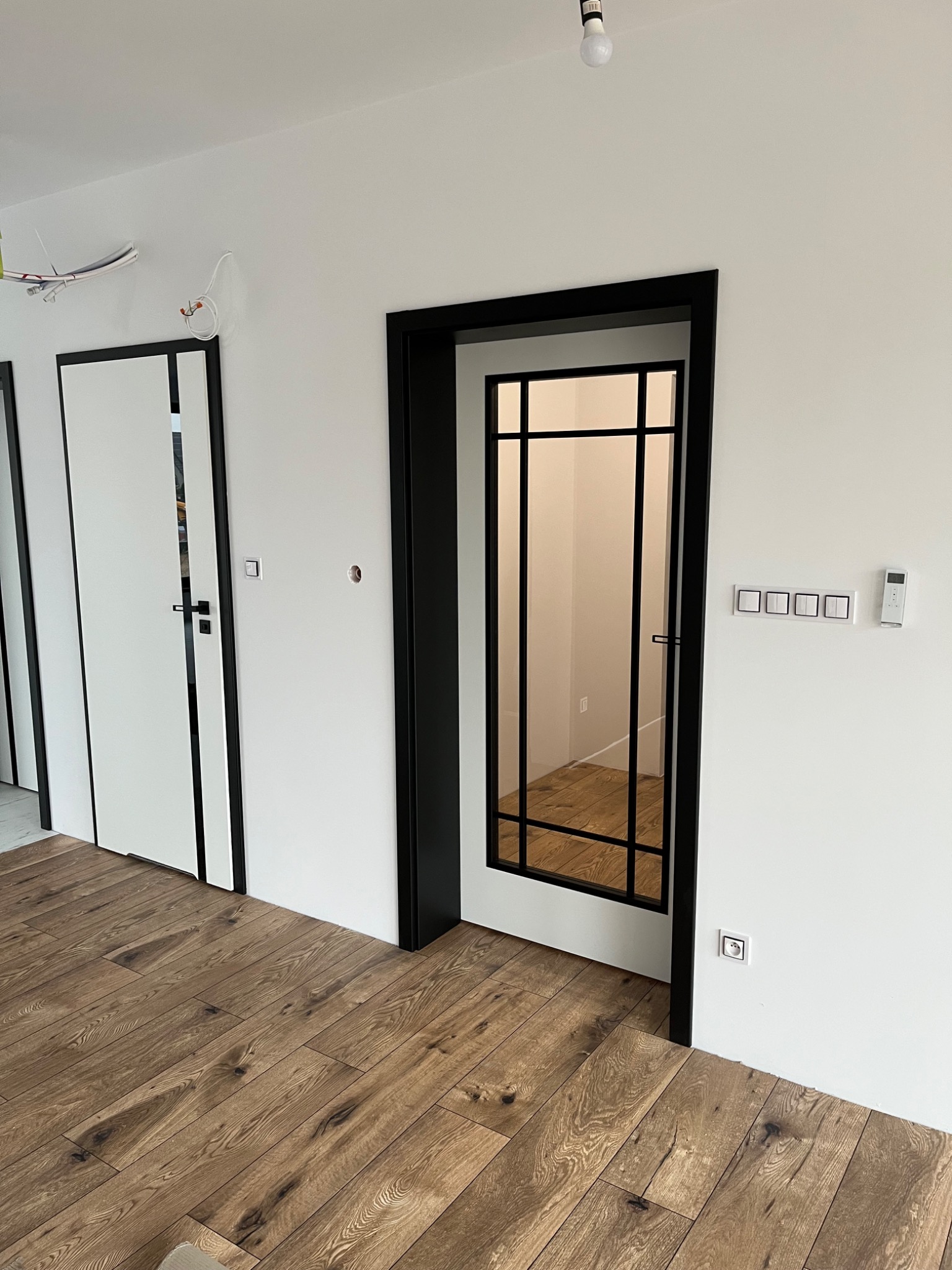 Nowoczesne, białe drzwi wewnętrzne z czarną ramą i szklanymi panelami, widoczne na tle drewnianej podłogi i białych ścian w minimalistycznym wnętrzu.