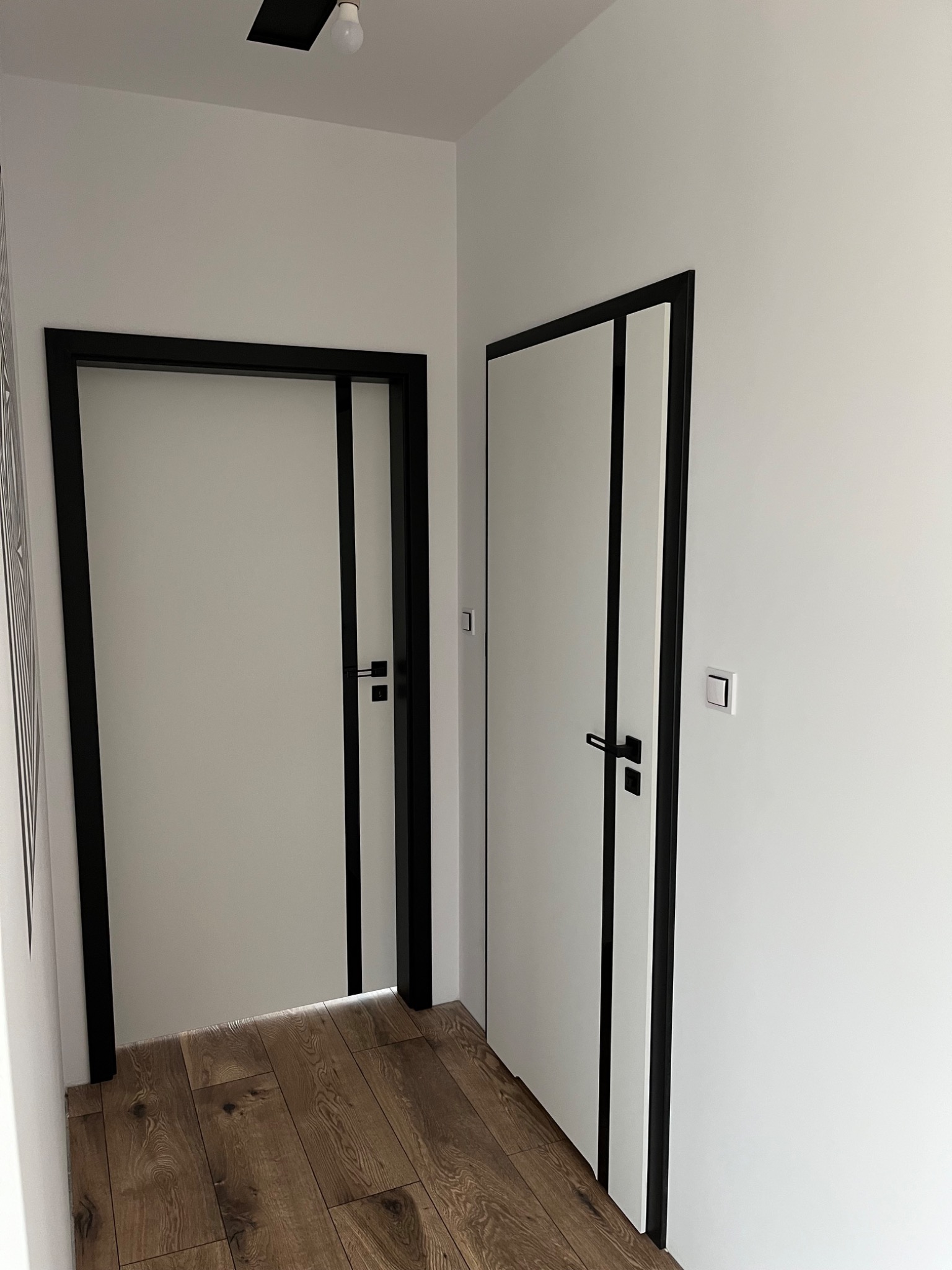 Nowoczesne, białe skrzydła drzwiowe z czarnymi ościeżnicami w jasnym wnętrzu, widok na dwa wejścia z drewnianą podłogą. Minimalistyczny design.