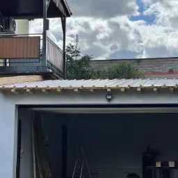 Nowo wykończony dach garażu z blachy trapezowej, widoczna listwa okapowa i oświetlenie zewnętrzne, w tle balkon domu i pochmurne niebo.