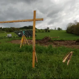 Prace przygotowawcze pod instalację paneli słonecznych: niwelator na statywie, drewniane konstrukcje i wytyczony teren na tle zielonego wzgórza pod pochmurnym niebem.