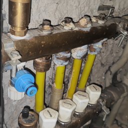 Dawid Litwinski - Zamknięty rozdzielacz instalacji grzewczej z żółtymi rurami, widoczny na tle surowej ściany. Elementy instalacji noszą ślady użytkowania.