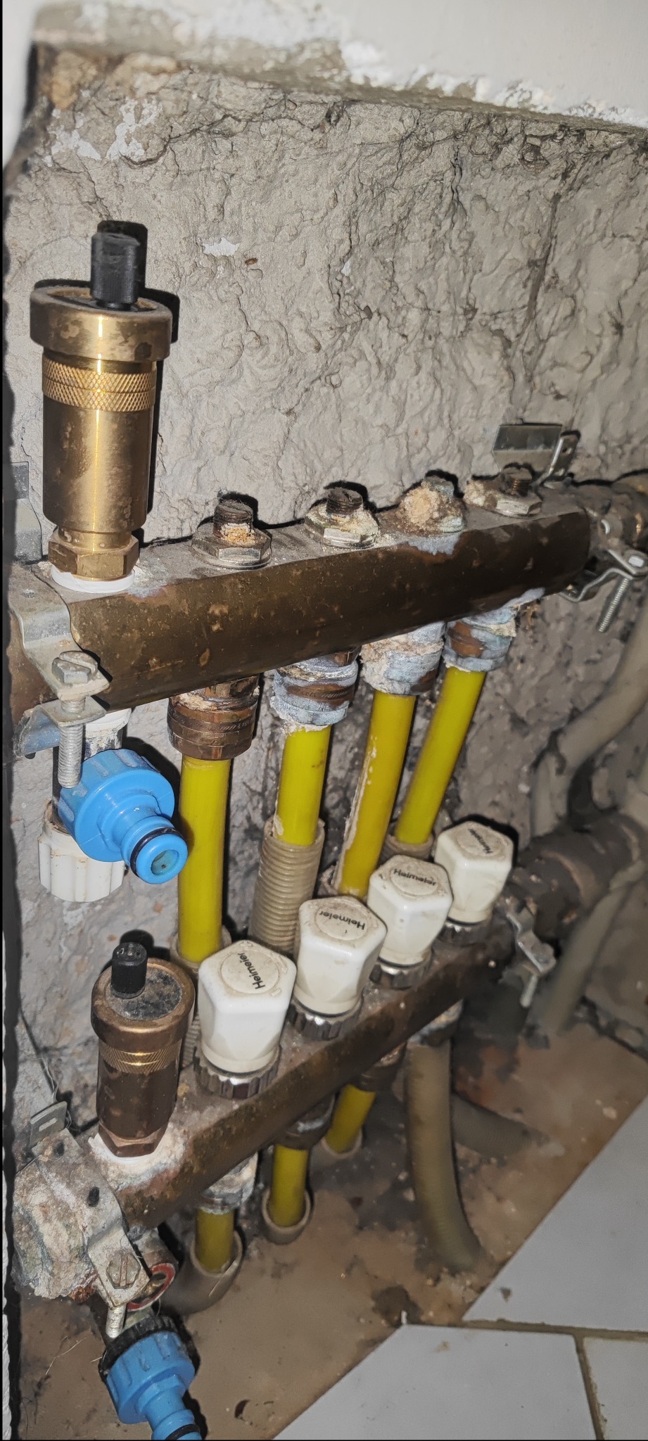 Zamknięty rozdzielacz instalacji grzewczej z żółtymi rurami, widoczny na tle surowej ściany. Elementy instalacji noszą ślady użytkowania.