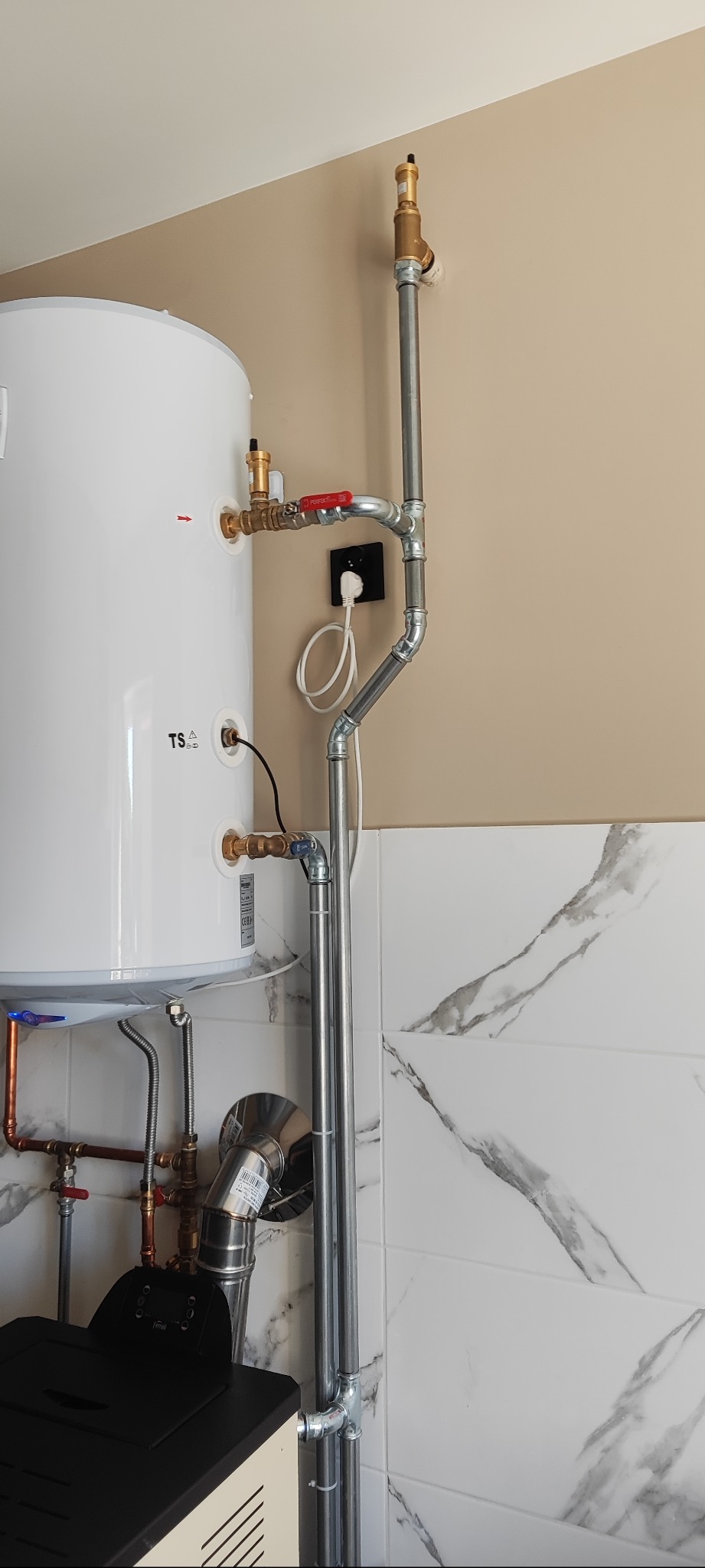 Biały termo kominek na pellet Ferroli z podłączonymi rurami i instalacją, zamontowany przy ścianie z marmurowymi płytkami. Widoczny fragment kotła i instalacja elektryczna.