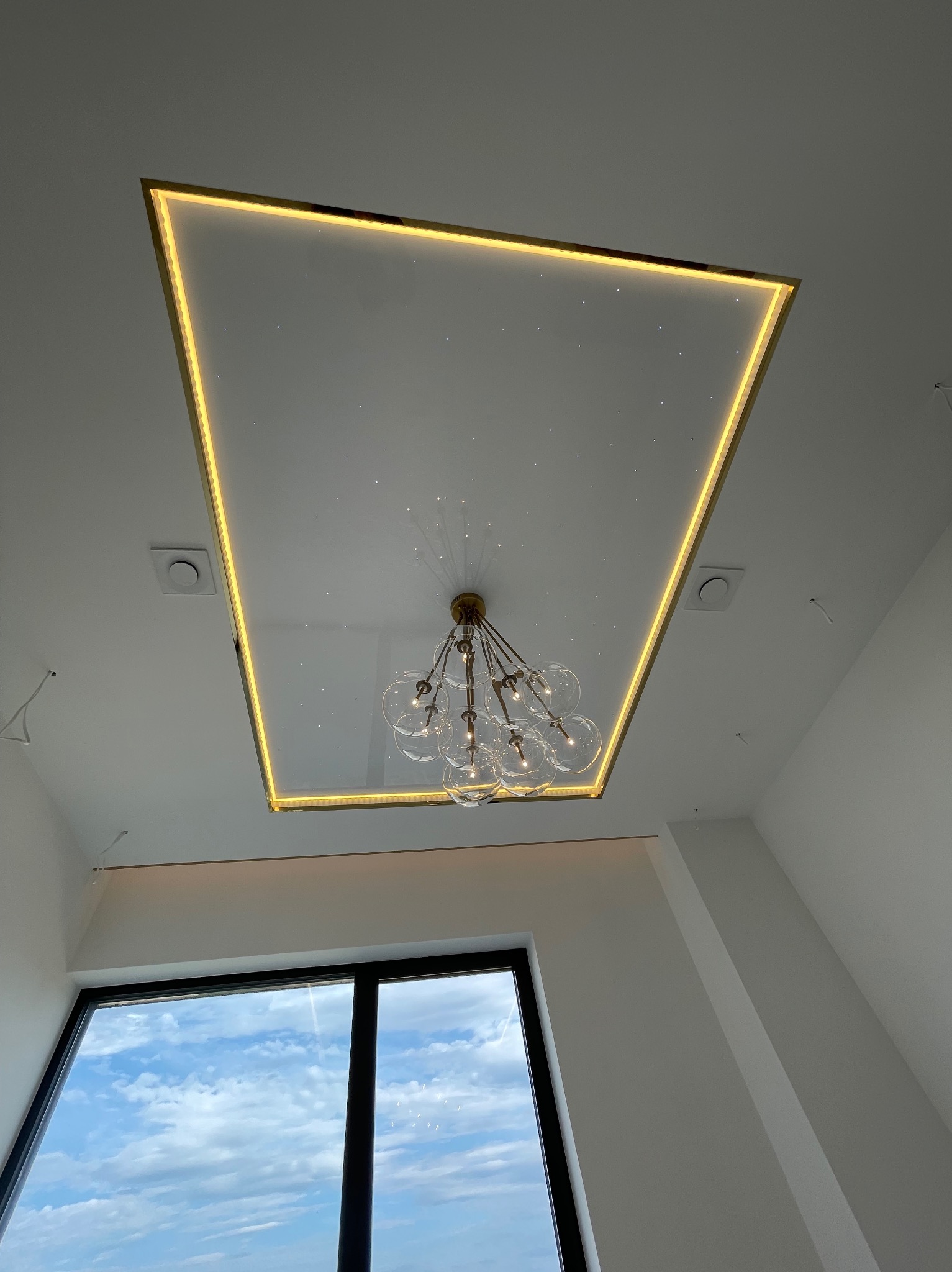 Biały sufit z efektem gwiazd, obramowany złotą ramą z oświetleniem LED, w centrum designerski żyrandol z przezroczystymi kulami, widok z dołu.