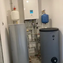 Kompaktowa kotłownia z dwoma zbiornikami akumulacyjnymi ciepła, białym kotłem Panasonic z wyświetlaczem i rozbudowaną instalacją rurową w małym pomieszczeniu z wzorzystą podłogą.