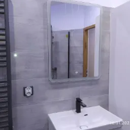 Nowoczesna łazienka z szarymi płytkami, podświetlanym lustrem, białą umywalką z czarną armaturą i grzejnikiem drabinkowym w kolorze antracytu.