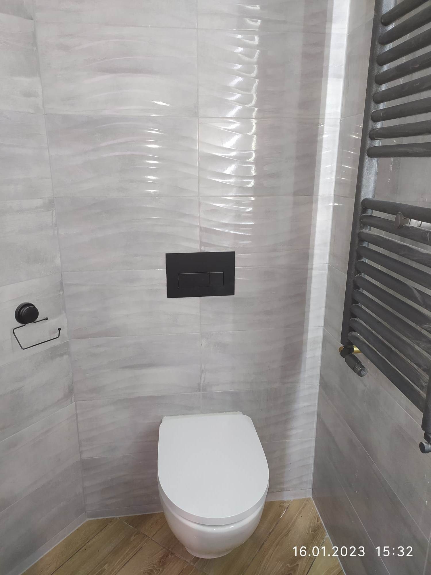 Nowoczesna łazienka z białą miską WC, szarymi płytkami o falistej strukturze, czarnym przyciskiem spłuczki, grzejnikiem drabinkowym w kolorze antracytu i podłogą imitującą drewno.