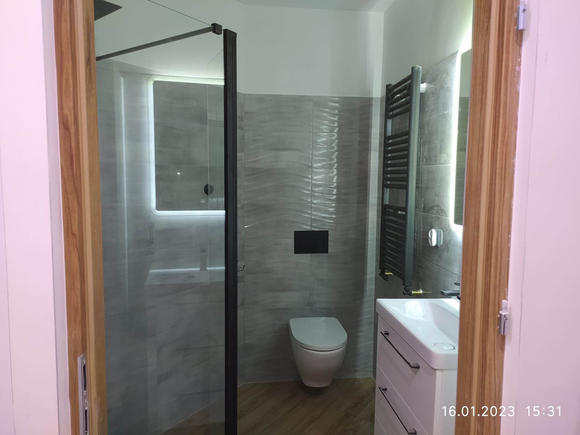 Wykończona łazienka z kabiną prysznicową typu walk-in z czarnymi profilami, wiszącą toaletą, grzejnikiem drabinkowym i umywalką z szafką w odcieniach bieli i szarości z płytkami imitującymi kamień.