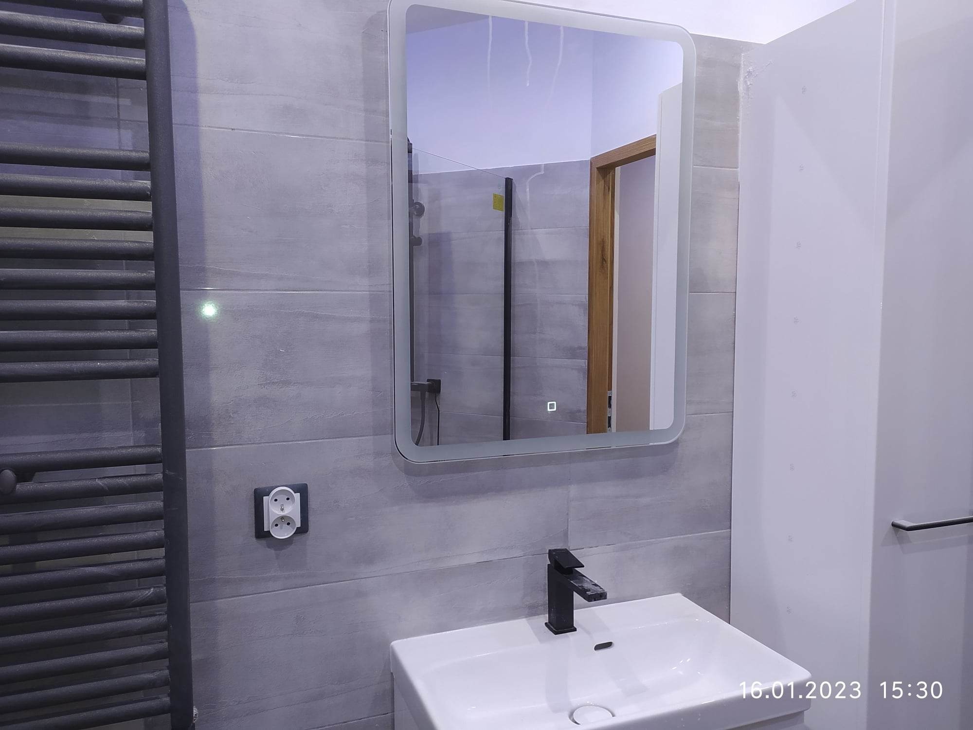 Nowoczesna łazienka z szarymi płytkami, podświetlanym lustrem, białą umywalką z czarną armaturą i grzejnikiem drabinkowym w kolorze antracytu.