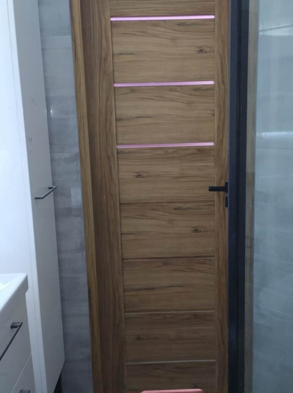 Drzwi łazienkowe z drewna z poziomymi, podświetlonymi paskami, widoczne obok umywalki i kabiny prysznicowej.