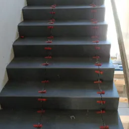 Schody wyłożone szarymi płytkami, z użyciem czerwonych klipsów poziomujących, widoczne ślady po kleju, w trakcie prac wykończeniowych.