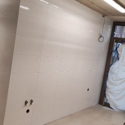 ART-BUD - Białe, błyszczące płytki ceramiczne ułożone na ścianach w trakcie remontu łazienki; widoczne przygotowane otwory pod armaturę i zabezpieczone folią drzwi.
