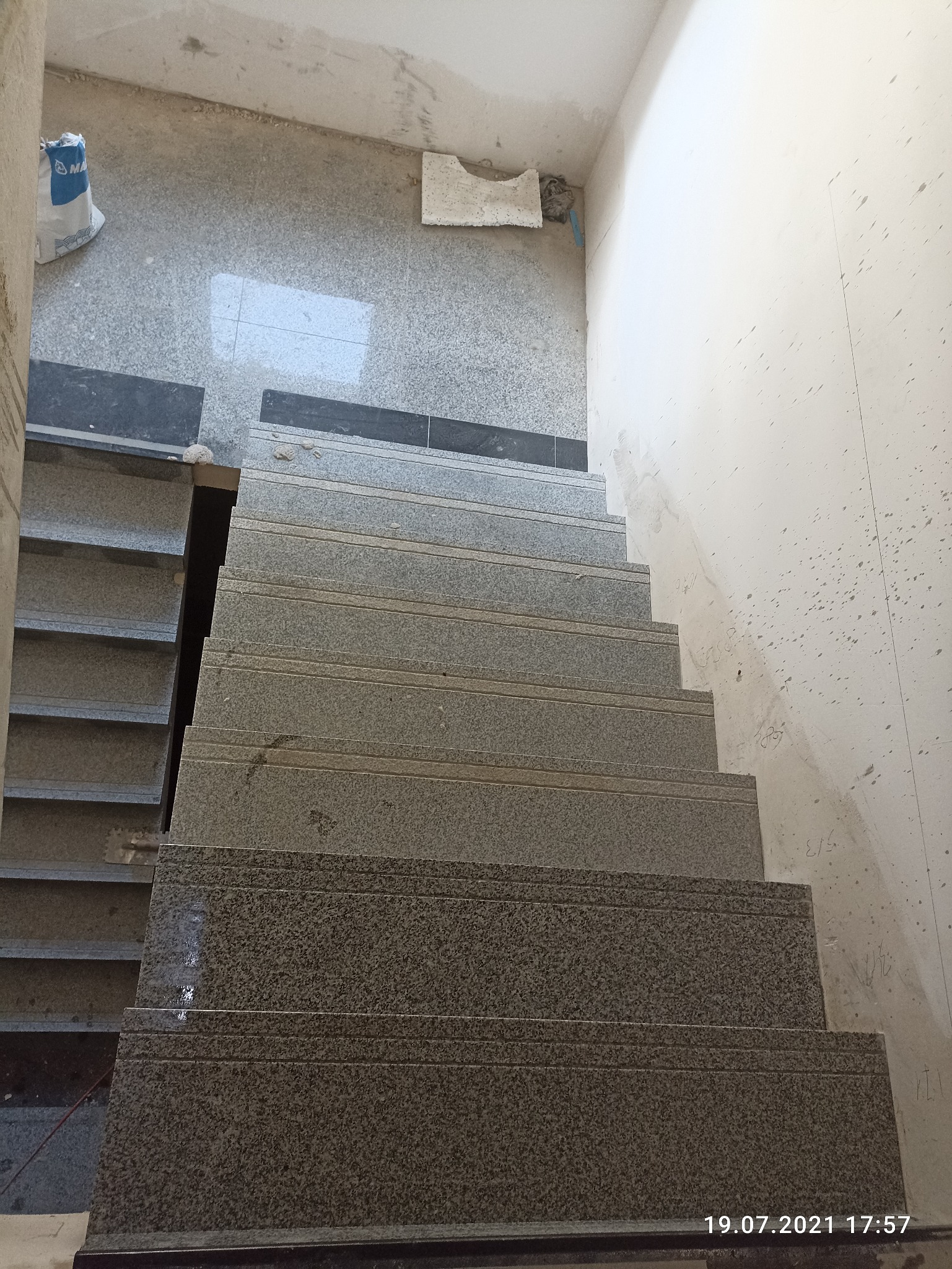 Klatka schodowa w trakcie wykańczania, widoczne szare płytki na schodach, surowy beton na ścianach i podłodze, worek z zaprawą w rogu.