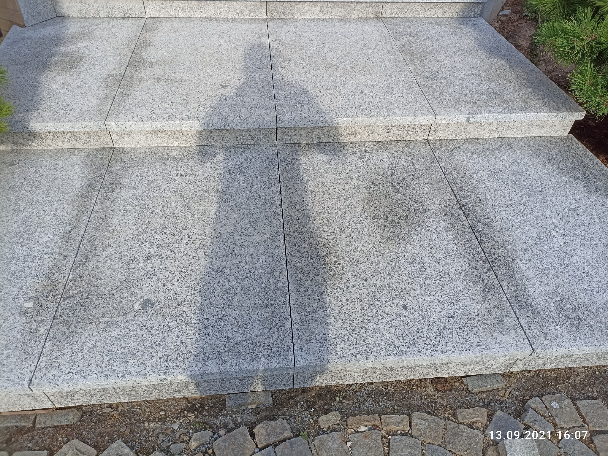 Granitowe schody zewnętrzne prowadzące do domu, cień sylwetki osoby na stopniach, obrzeże z kostki brukowej.