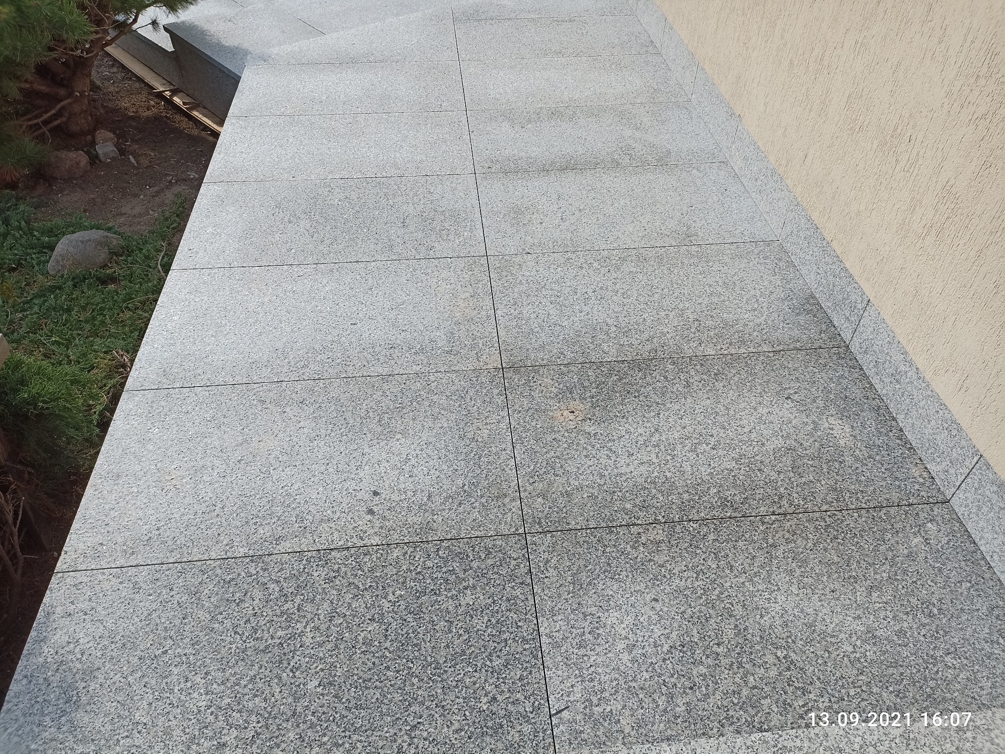 Granitowe płyty chodnikowe ułożone na zewnątrz budynku, widoczne z bliska, z fragmentem trawnika i drzewa po lewej stronie oraz fragmentem ściany budynku po prawej stronie. Płyty o szorstkiej...