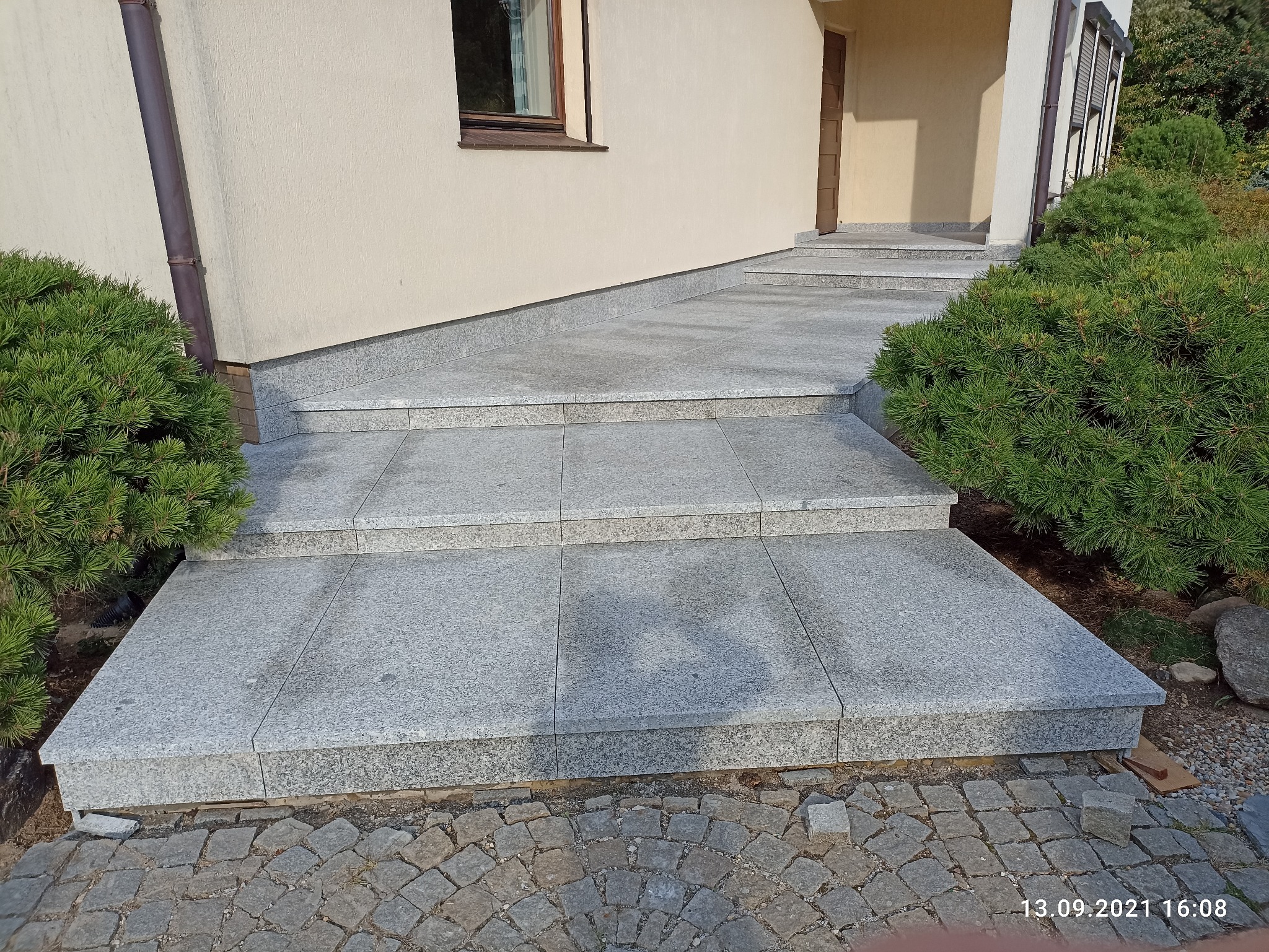 Granitowe schody wejściowe do domu, wykończone szarą kostką brukową, z zielonymi krzewami po bokach