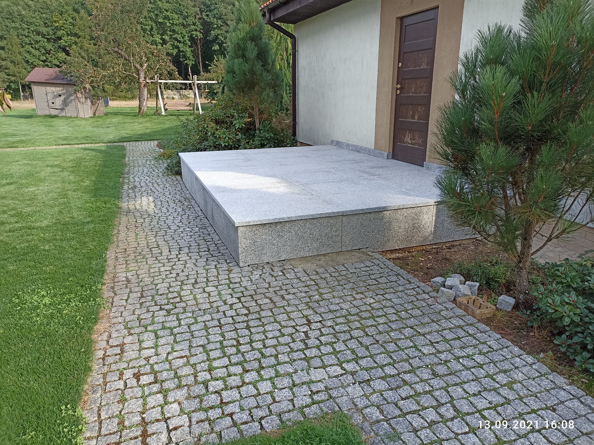 Wykończony, granitowy podest przed wejściem do budynku, z kostką brukową prowadzącą do niego, widok z poziomu gruntu, w tle trawnik z huśtawką i domkiem narzędziowym.