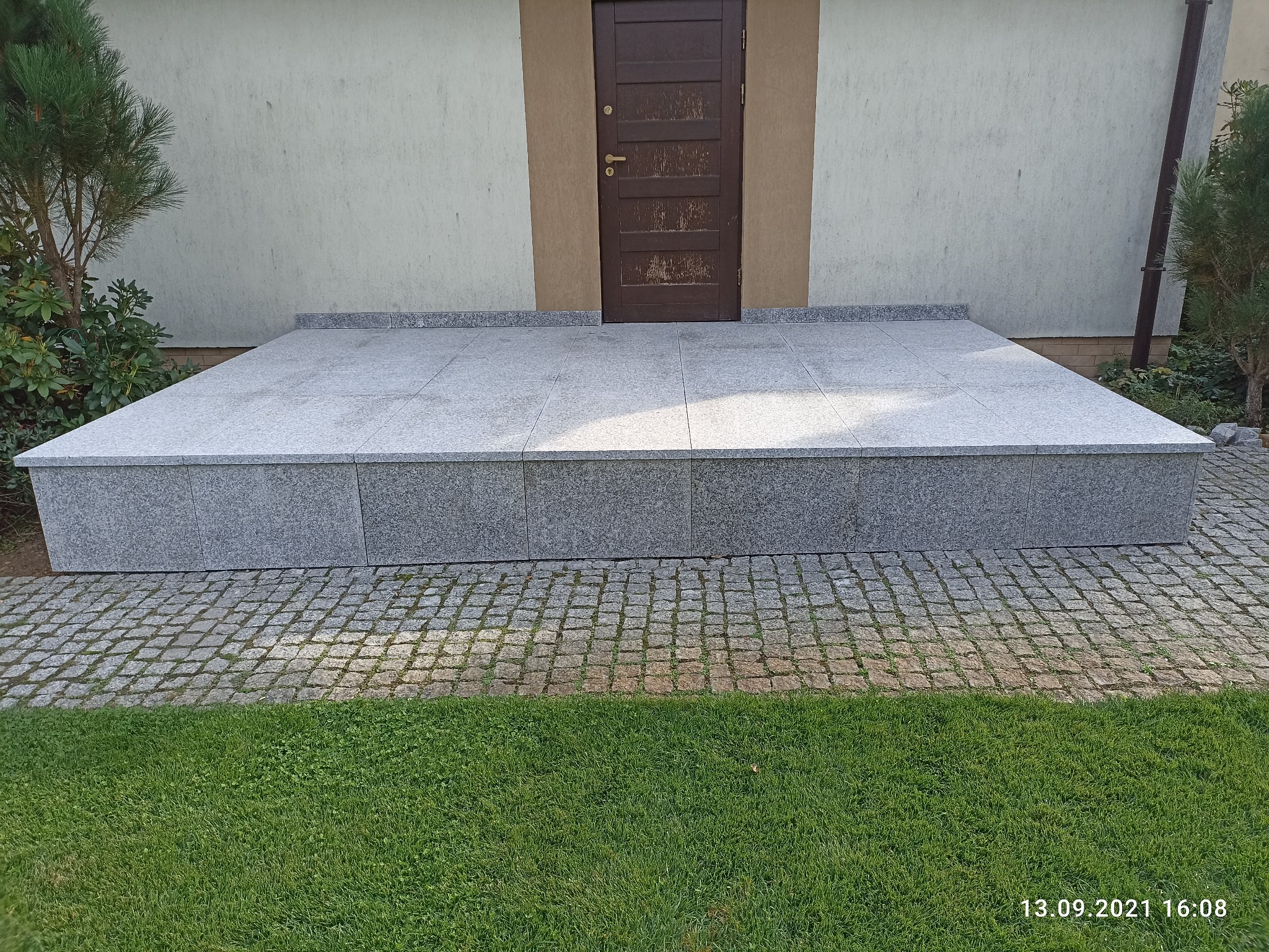 Granitowy podest wejściowy z czterema stopniami, wyłożony płytami i obłożony kostką brukową, prowadzący do brązowych drzwi wejściowych.