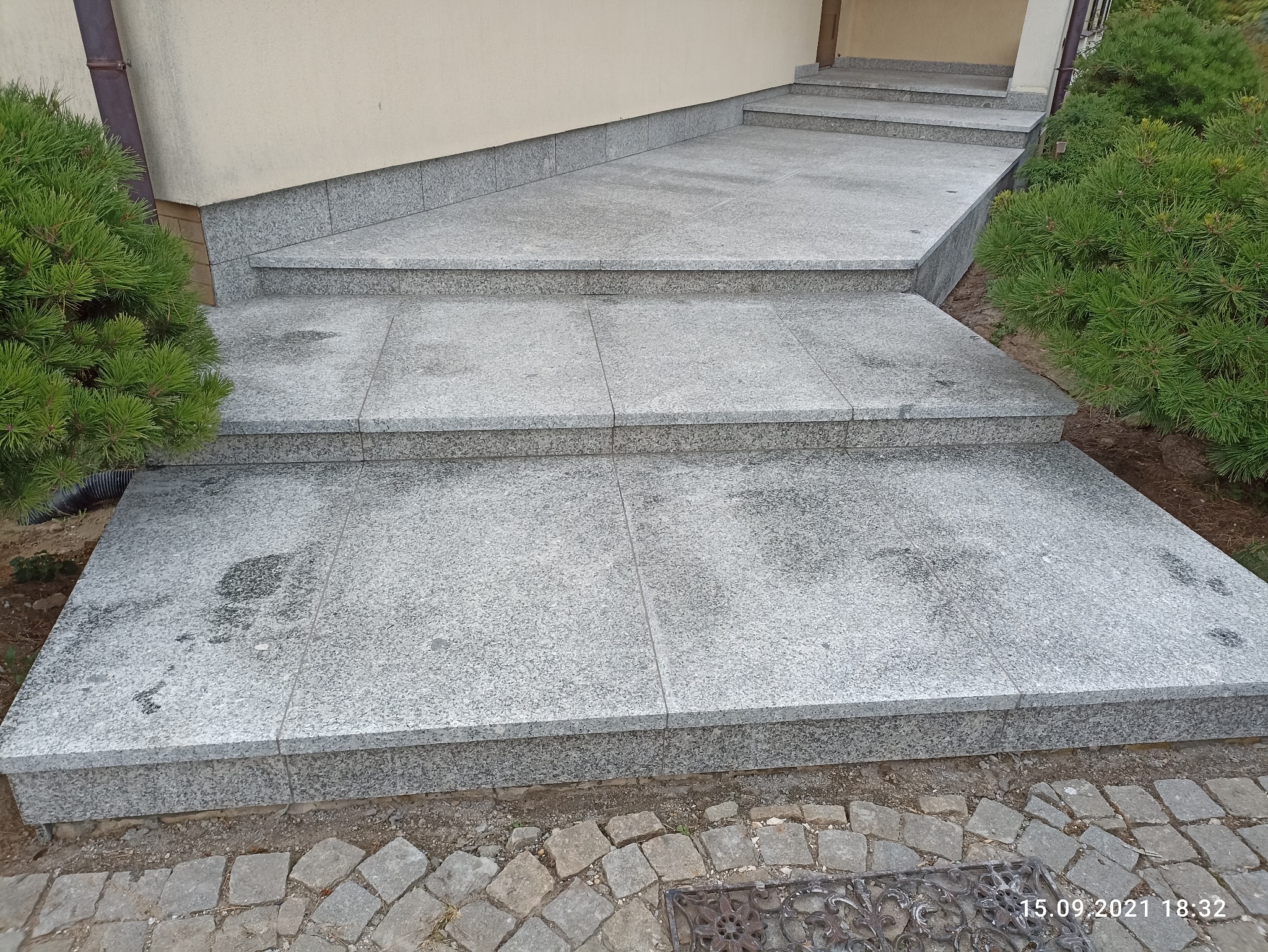 Schody wejściowe wyłożone szarymi płytami granitowymi, z widocznymi spoinami, obok iglaki, widoczny fragment chodnika z kostki brukowej.