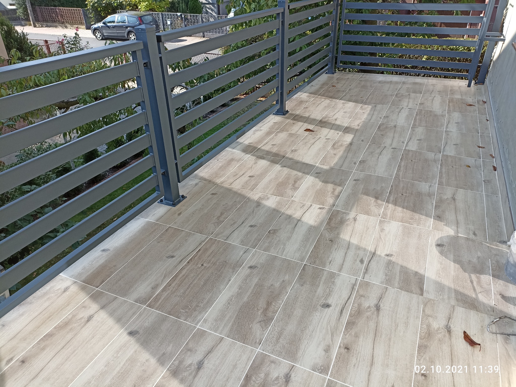 Wyłożony płytkami balkon z szarą metalową balustradą. Płytki imitują drewno i są ułożone w kwadraty z jasnymi fugami. Na podłodze leżą pojedyncze, uschnięte liście.