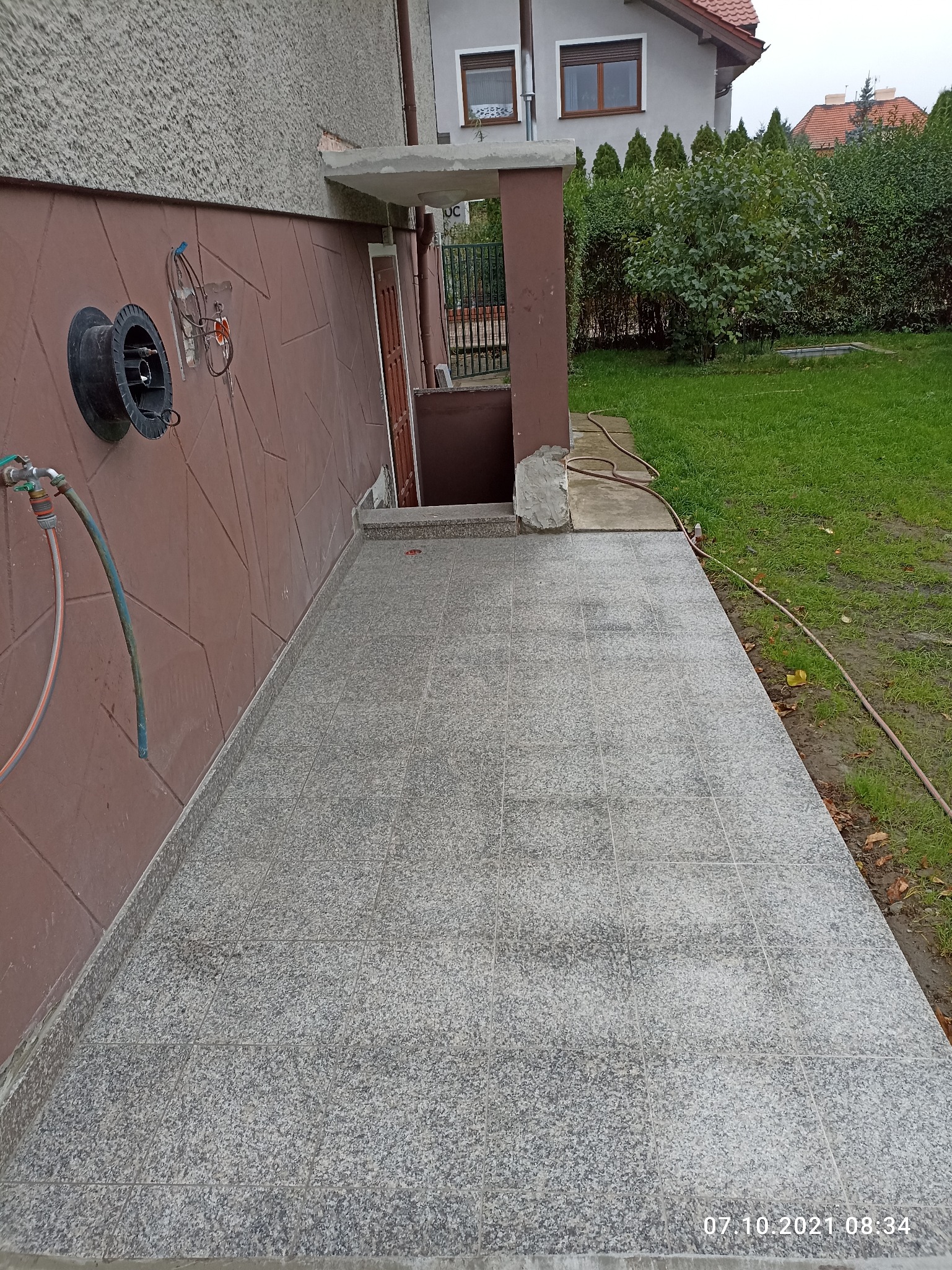Wykończony taras przed domem z szarych płytek, widoczny kran z wężem i niezabezpieczone przewody elektryczne na ścianie.