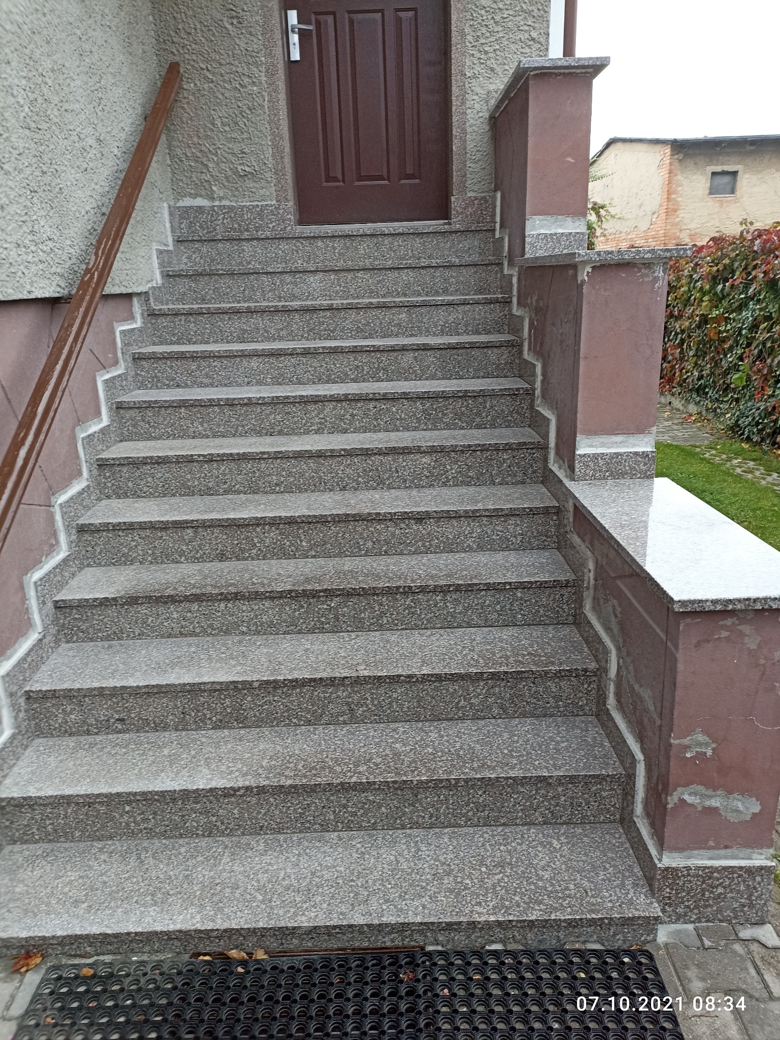 Zewnętrzne schody wejściowe wyłożone szarym kamieniem z brązowymi balustradami, prowadzące do brązowych drzwi wejściowych.