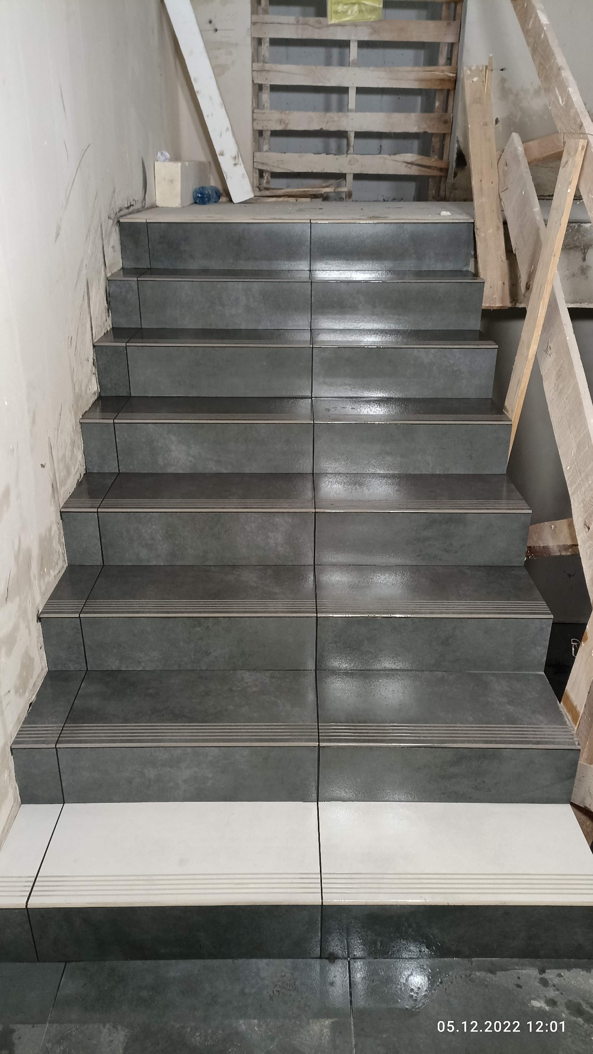 Klatka schodowa w trakcie wykańczania, z szarymi płytkami na stopniach i białymi na podeście, widoczne zabezpieczenia budowlane i data wykonania zdjęcia