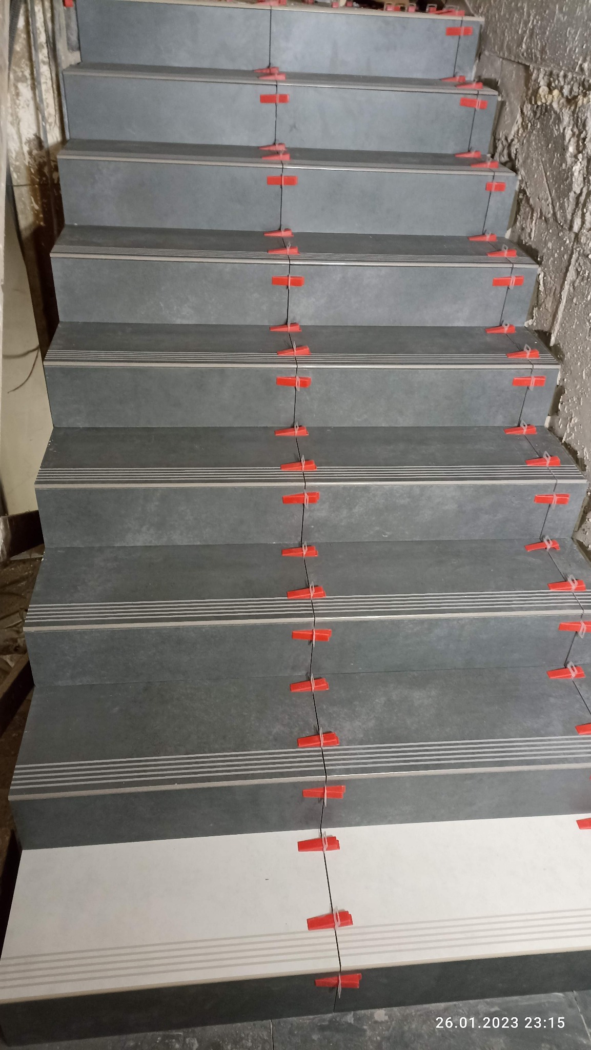 Schody wyłożone szarymi płytkami z poziomymi paskami, z użyciem czerwonych krzyżyków dystansowych, na etapie montażu.