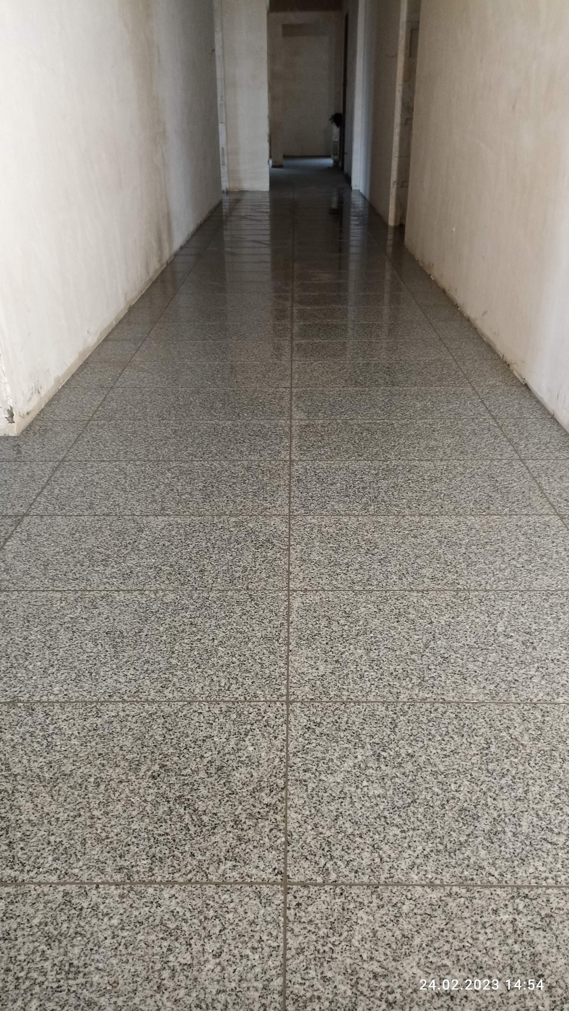 Korytarz z nowo położoną, błyszczącą, szarą podłogą z płytek imitujących granit, ściany surowe, w tle widoczne kolejne pomieszczenia w trakcie remontu.