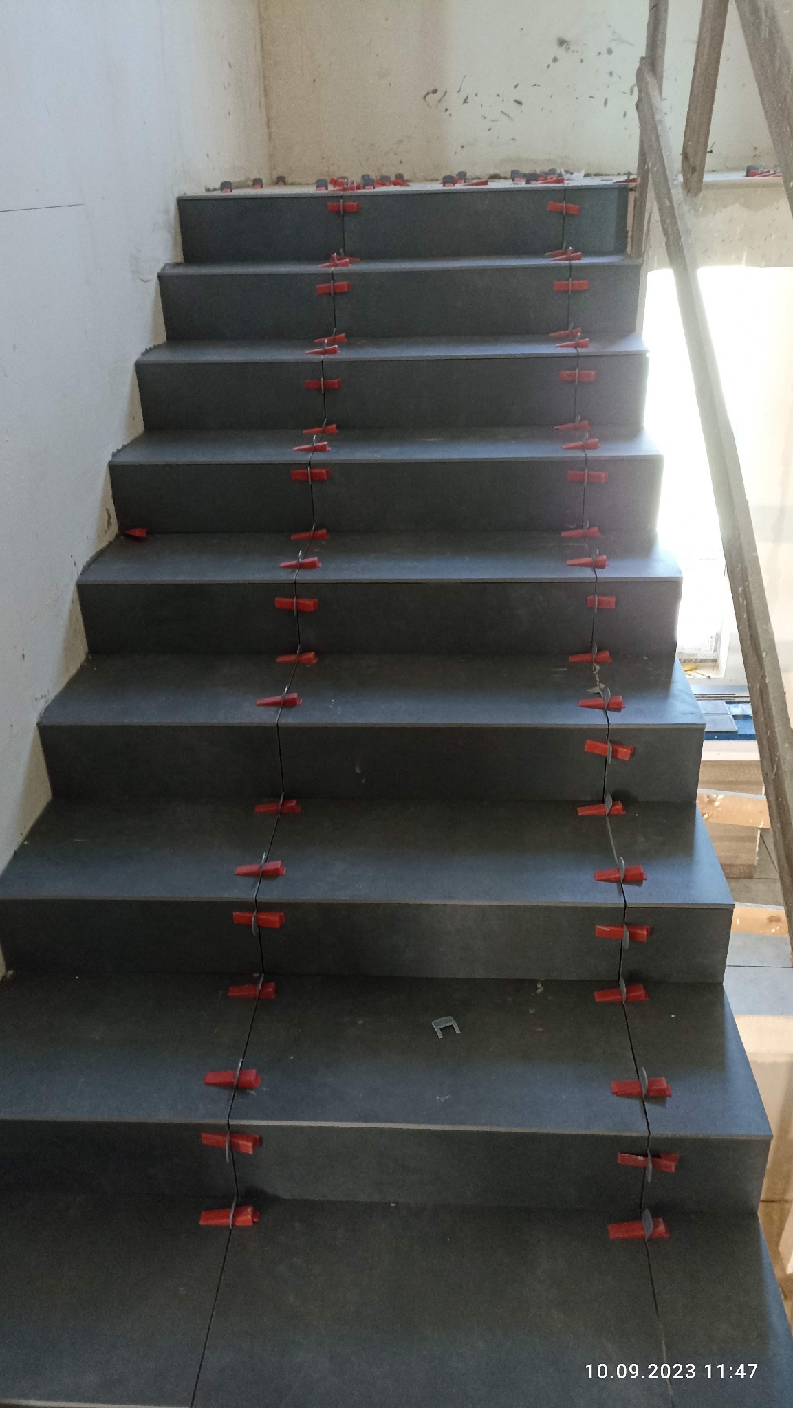 Schody wyłożone szarymi płytkami, z użyciem czerwonych klipsów poziomujących, widoczne ślady po kleju, w trakcie prac wykończeniowych.