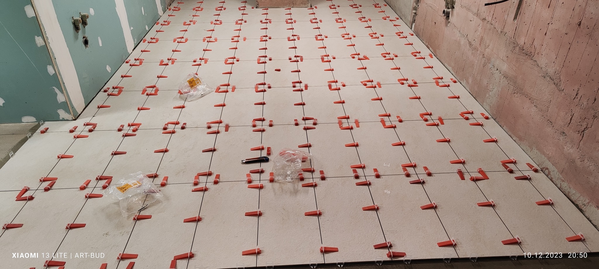 Układanie jasnych płytek podłogowych z użyciem systemu poziomowania, widoczne krzyżyki dystansowe, paczka z klinami i nóż introligatorski na powierzchni, ściany w surowym stanie przed wykończeniem.