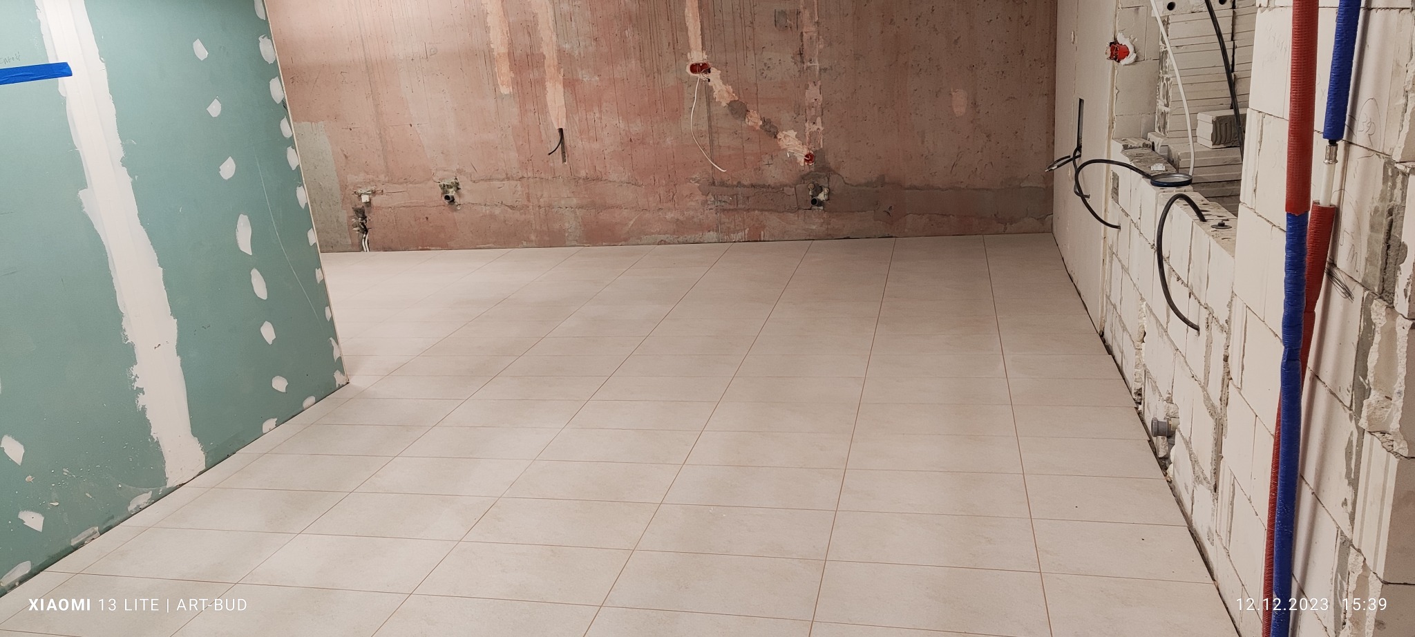 Wnętrze w trakcie remontu: ułożone jasne płytki podłogowe, surowe ściany z widocznymi instalacjami elektrycznymi i hydraulicznymi, fragment ściany z płyt gipsowo-kartonowych z widocznymi punktami...