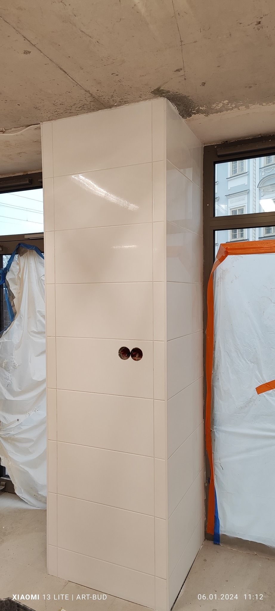 Pionowa kolumna obłożona białymi, błyszczącymi płytkami w trakcie prac wykończeniowych, widoczne dwa otwory na gniazdka elektryczne, okno z widokiem na budynek i folia ochronna na ścianie.