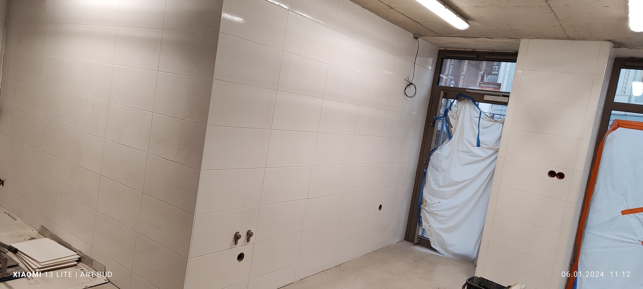 Białe, błyszczące płytki ceramiczne ułożone na ścianach w trakcie remontu łazienki; widoczne przygotowane otwory pod armaturę i zabezpieczone folią drzwi.