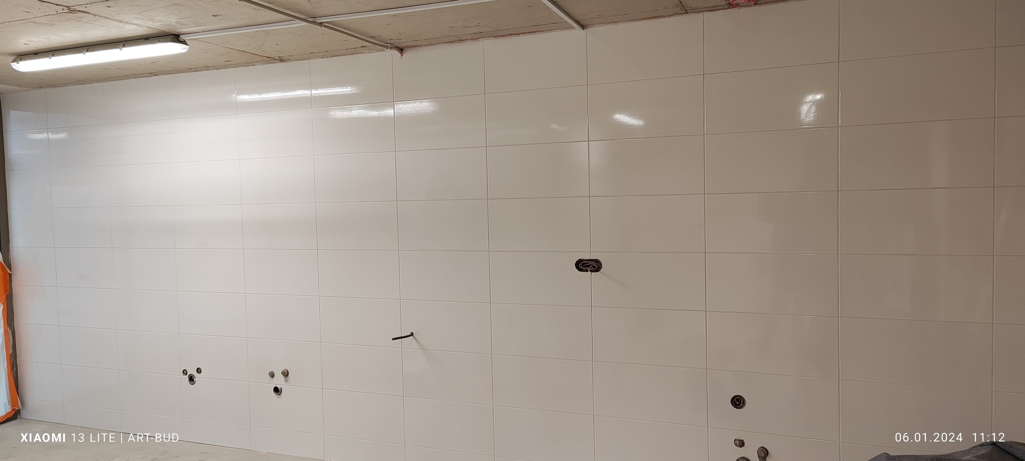 Ściana w trakcie układania płytek ceramicznych w jasnym odcieniu, widoczne otwory na instalacje wodne i elektryczne, surowy betonowy sufit z lampą i instalacją elektryczną.