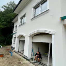 Biała elewacja domu jednorodzinnego z dekoracyjnymi obramowaniami okien i łukowym wejściem do garażu; pracownik w kucki odpoczywa w wejściu do garażu, widoczne nieukończone prace ziemne i materiały...