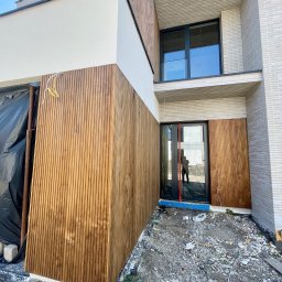 X-Y-BUD RAFAŁ ROMAŃSKI - Nowoczesny dom z elewacją z drewnianych paneli pionowych, połączoną z jasnym tynkiem i cegłą, widoczne przewody elektryczne, teren wokół domu w trakcie prac wykończeniowych.