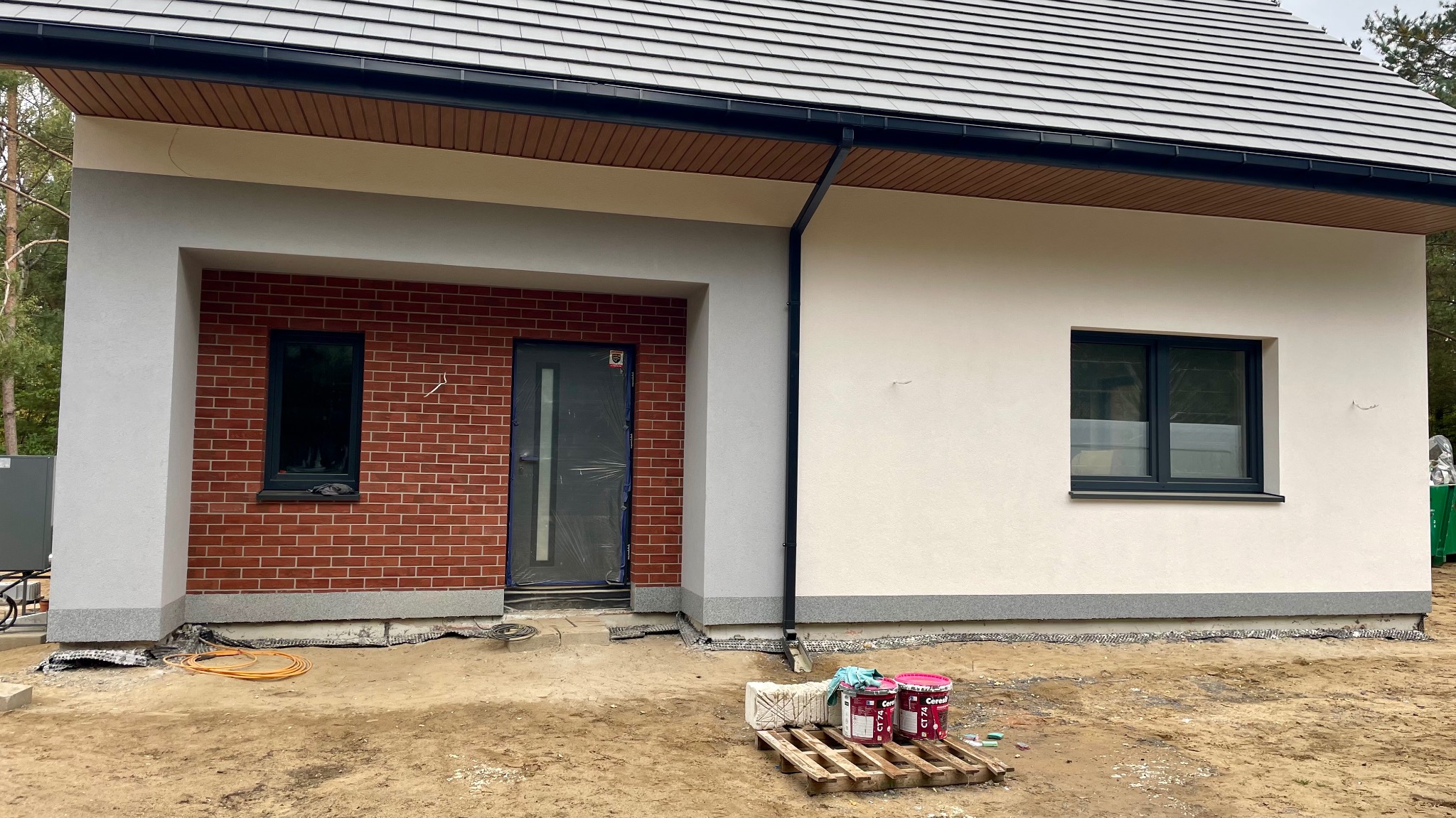 Nowoczesny dom jednorodzinny z elewacją w trakcie wykończenia: fragment ściany z białym tynkiem, fragment z czerwoną cegłą, okna w ciemnych ramach, puszki z farbą na palecie przed budynkiem.