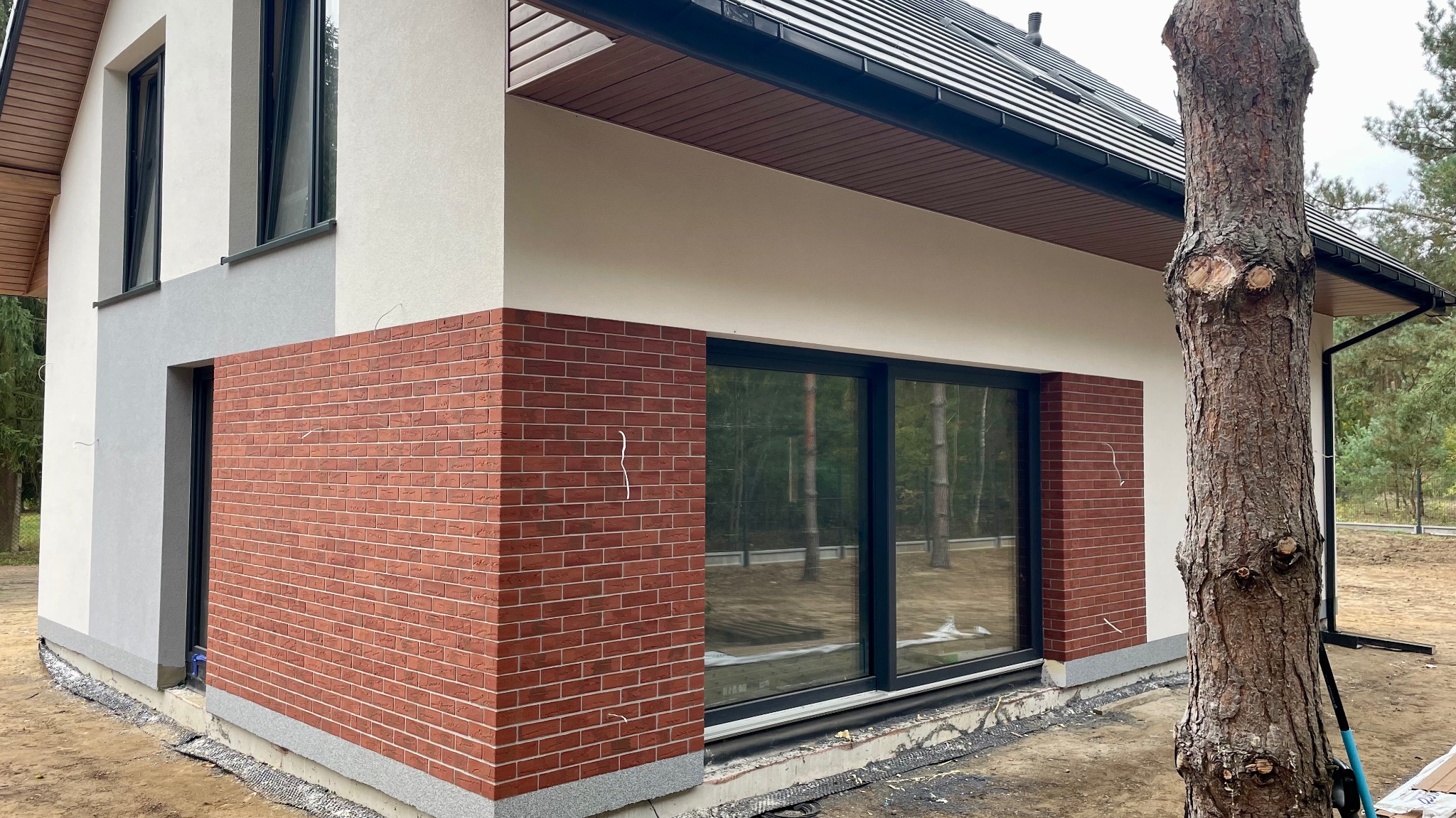 Fragment elewacji domu jednorodzinnego z oknem panoramicznym, wykończony częściowo cegłą klinkierową i jasnym tynkiem, widoczna instalacja elektryczna na ścianie oraz drzewo na pierwszym planie.
