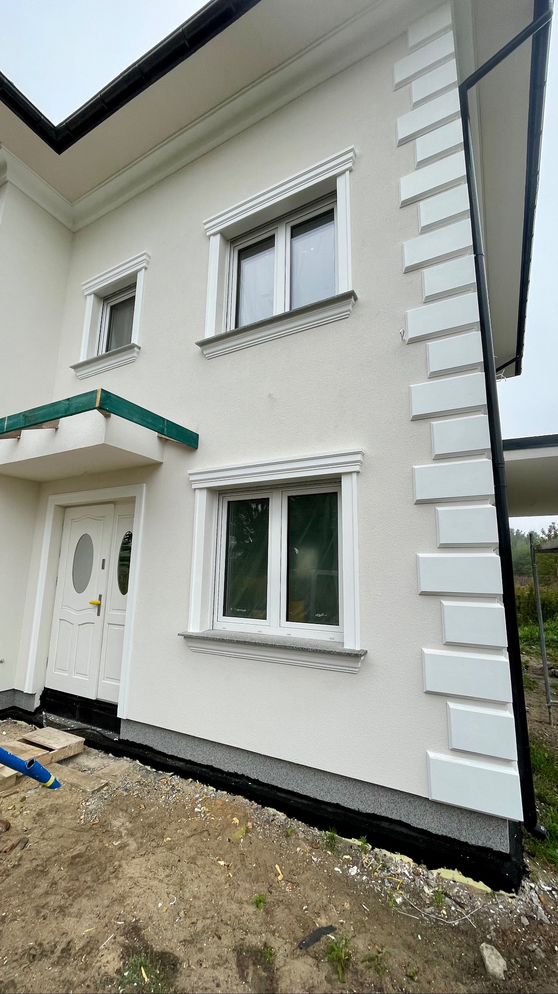 Nowoczesny dom jednorodzinny z białą elewacją, ozdobnymi elementami wokół okien i na narożniku, oraz widocznymi pracami wykończeniowymi przy gruncie.