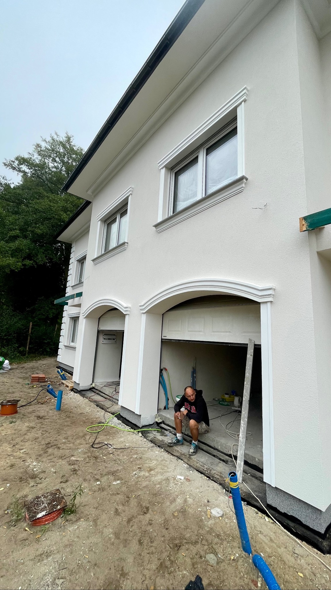 Biała elewacja domu jednorodzinnego z dekoracyjnymi obramowaniami okien i łukowym wejściem do garażu; pracownik w kucki odpoczywa w wejściu do garażu, widoczne nieukończone prace ziemne i materiały...