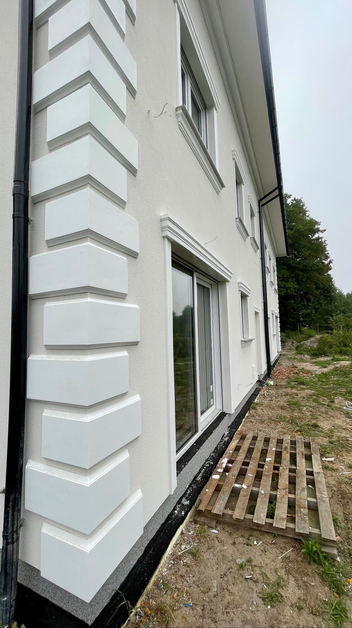 Fragment elewacji budynku z dekoracyjnymi, wystającymi elementami narożnymi i ozdobnymi obramowaniami okiennymi, widoczny czarny system rynnowy, paleta drewniana leżąca przy ścianie, w tle zieleń.