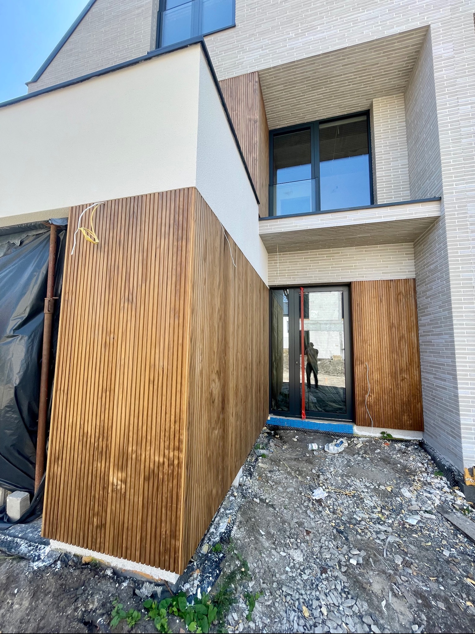 Nowoczesny dom z elewacją z drewnianych paneli pionowych, połączoną z jasnym tynkiem i cegłą, widoczne przewody elektryczne, teren wokół domu w trakcie prac wykończeniowych.