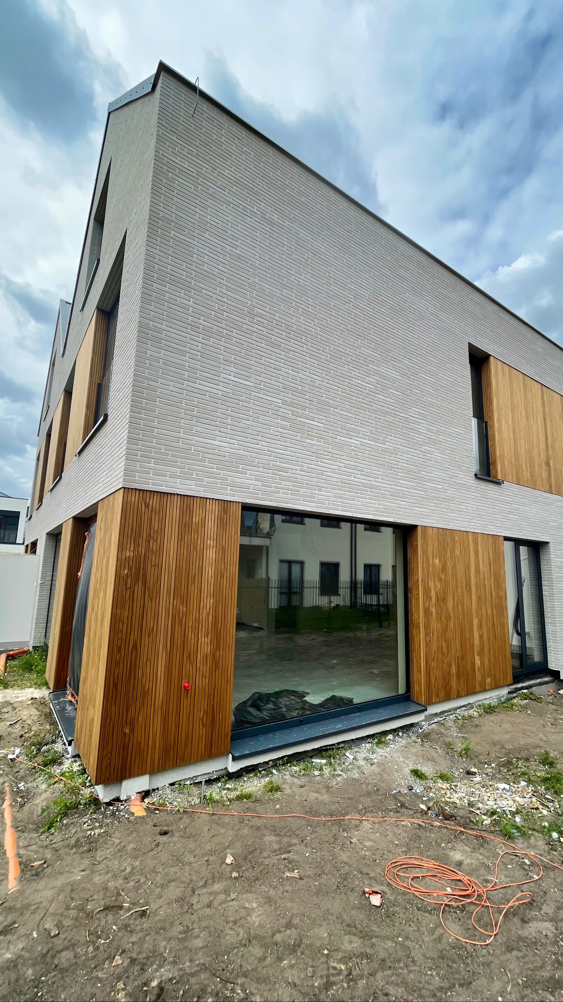 Nowoczesny dom z elewacją z jasnej cegły i drewnianymi panelami, widoczne okna i teren wokół budynku w trakcie prac wykończeniowych.