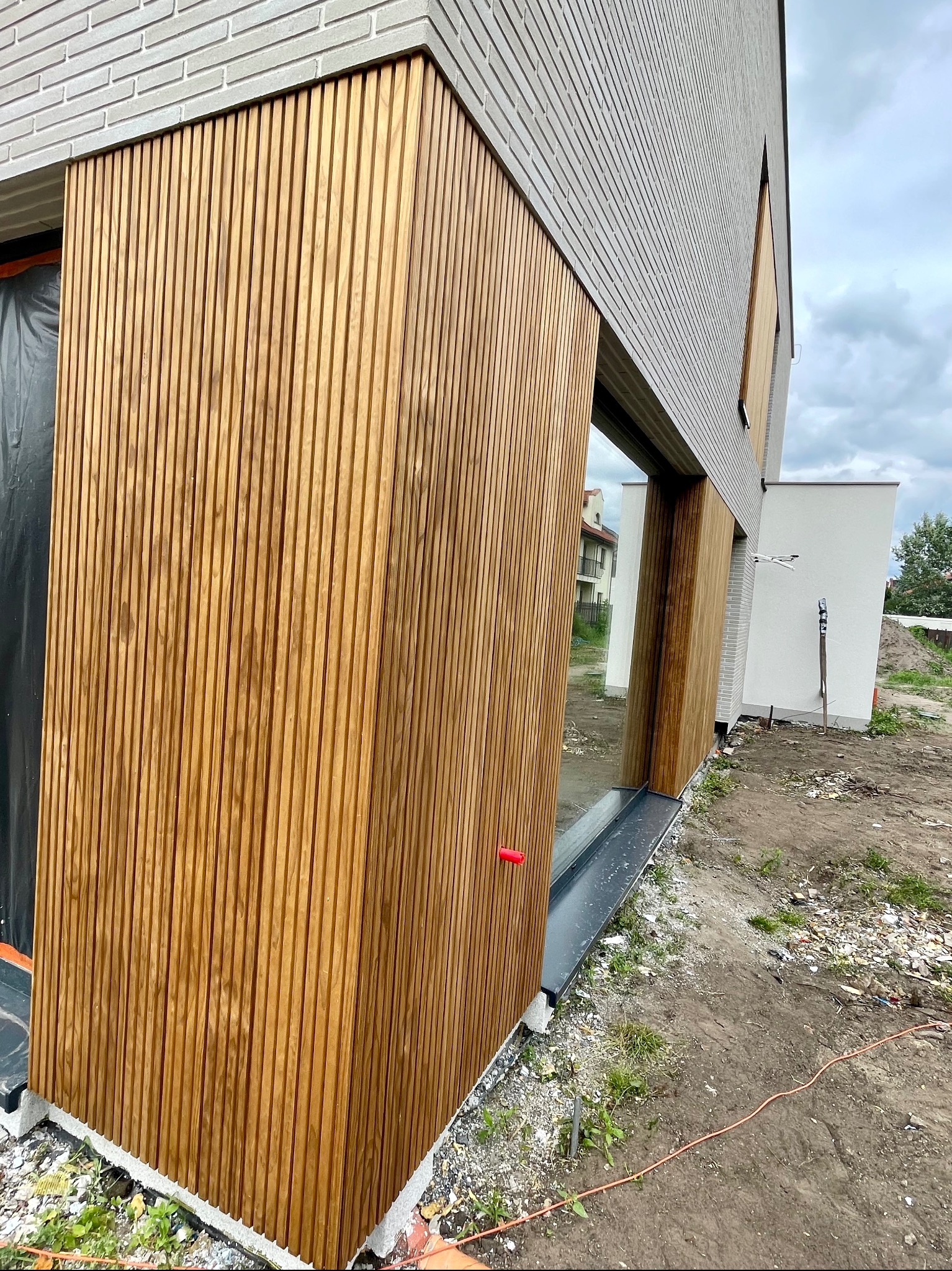 Nowoczesna elewacja budynku z szarej cegły i drewnianych paneli pionowych, widoczne okno z szarą ramą, teren wokół w trakcie prac wykończeniowych.