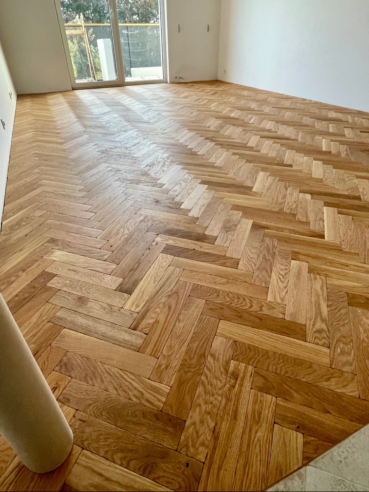 Ułożony parkiet w jodełkę francuską, jasne drewno, widok na okno balkonowe, świeżo po montażu, widoczne detale struktury drewna.