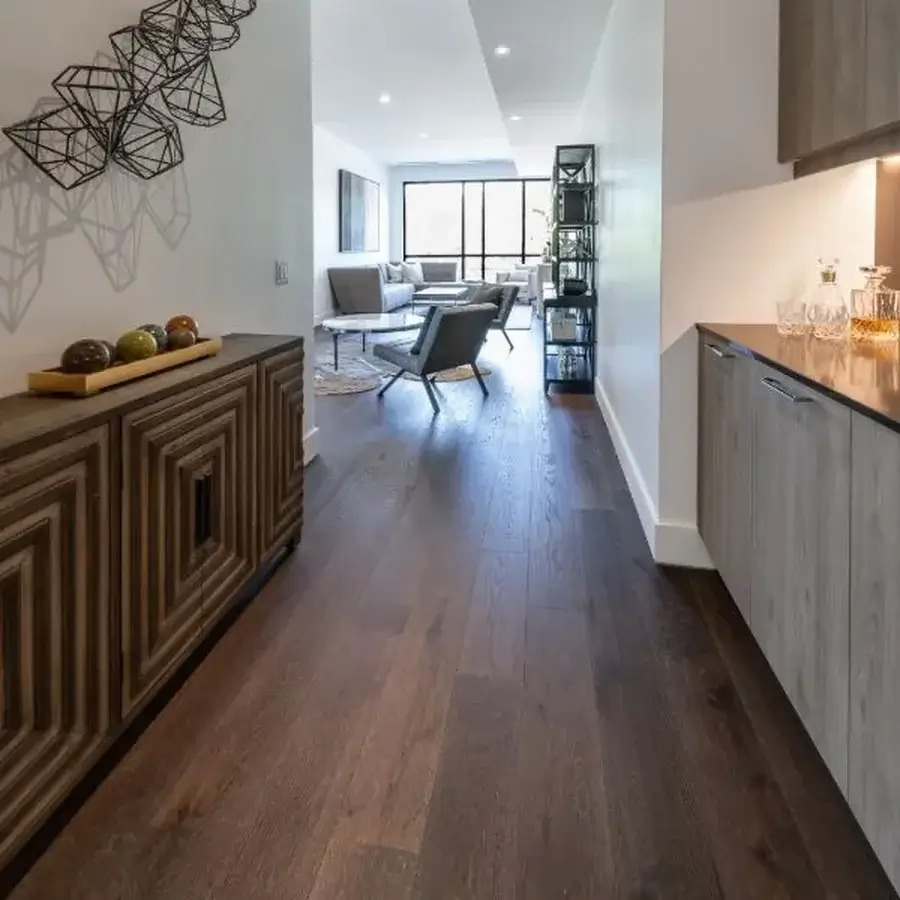 Ciemna, drewniana podłoga w korytarzu z widokiem na salon. Po lewej stronie ozdobna komoda, po prawej szafki kuchenne. Nowoczesny design wnętrza.