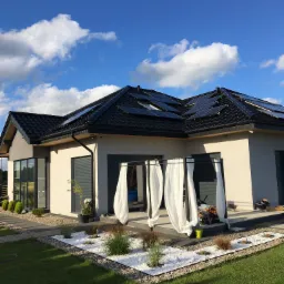 Nowoczesny dom jednorodzinny z ciemną dachówką i panelami słonecznymi, widok z ogrodu z białym kamieniem dekoracyjnym i zwiewnymi zasłonami na tarasie.