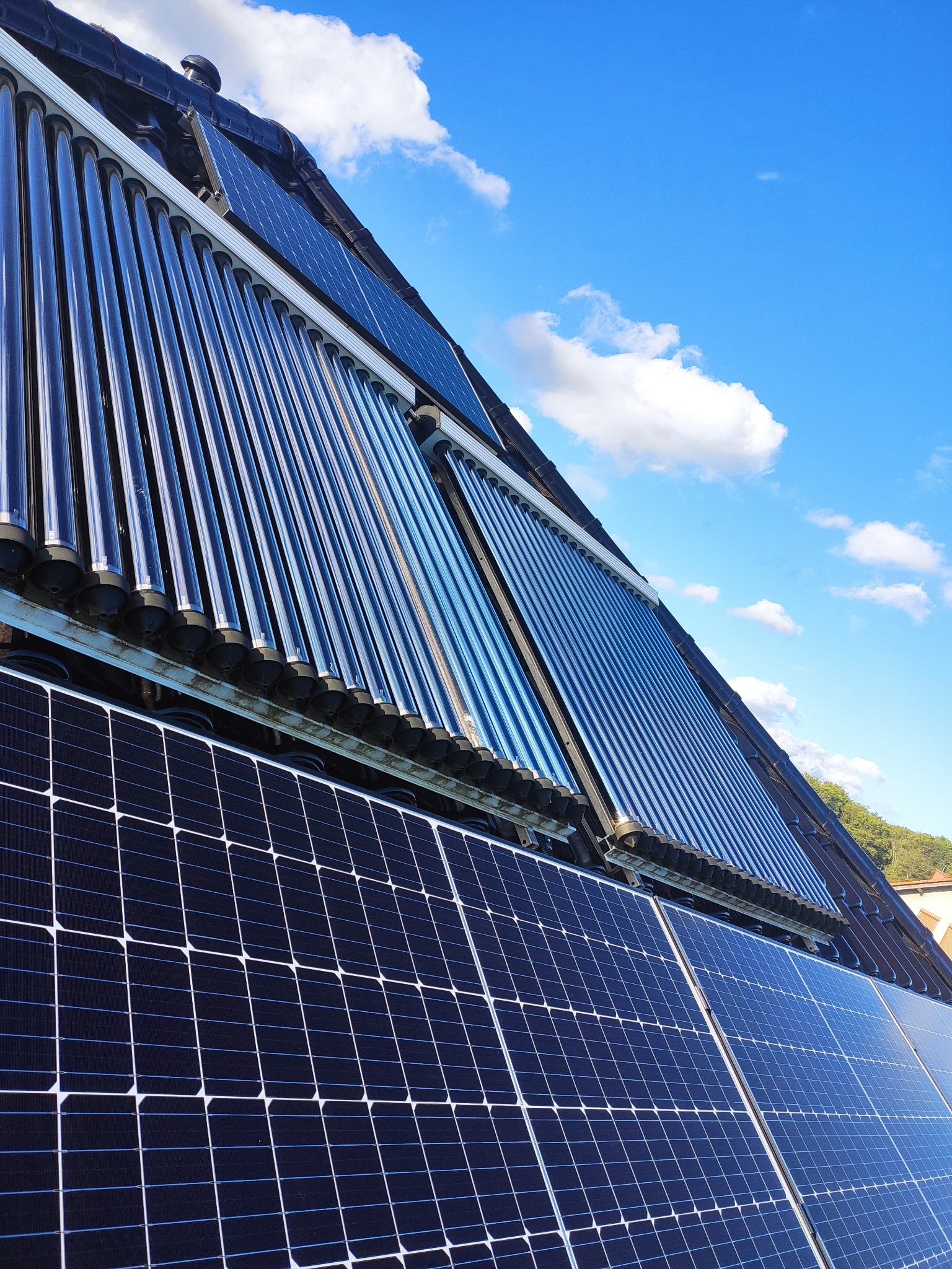 Panele fotowoltaiczne i kolektory słoneczne zamontowane na dachu z dachówki ceramicznej, widok z dołu na błękitne niebo z chmurami.