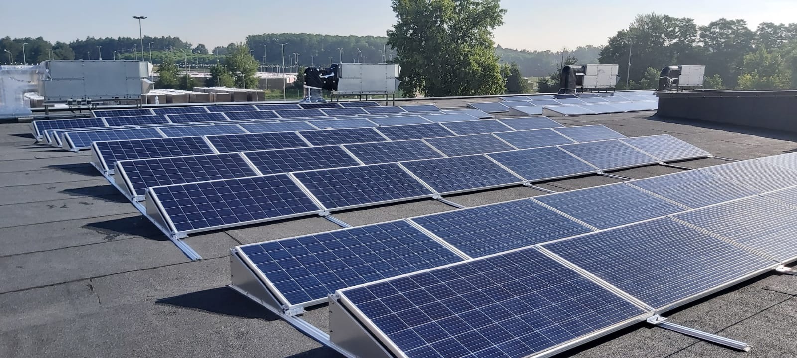 Rząd paneli fotowoltaicznych na płaskim dachu, z widocznymi elementami montażowymi i instalacją wentylacyjną w tle, oświetlonych jasnym, naturalnym światłem słonecznym.